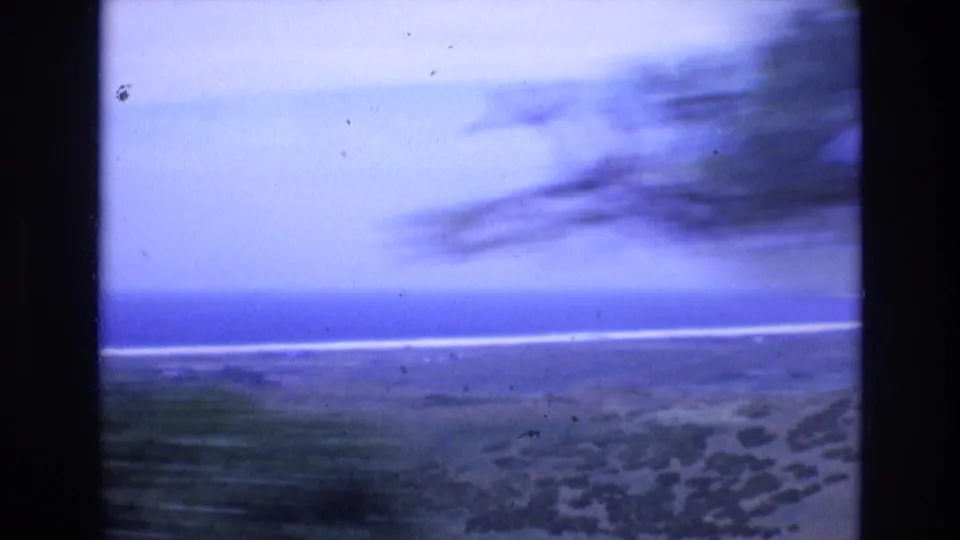 1972 Spain home-movie frame of a sandy shore with blurred dark vegetation or tree shapes, cloud bands, and a pale ocean horizon.