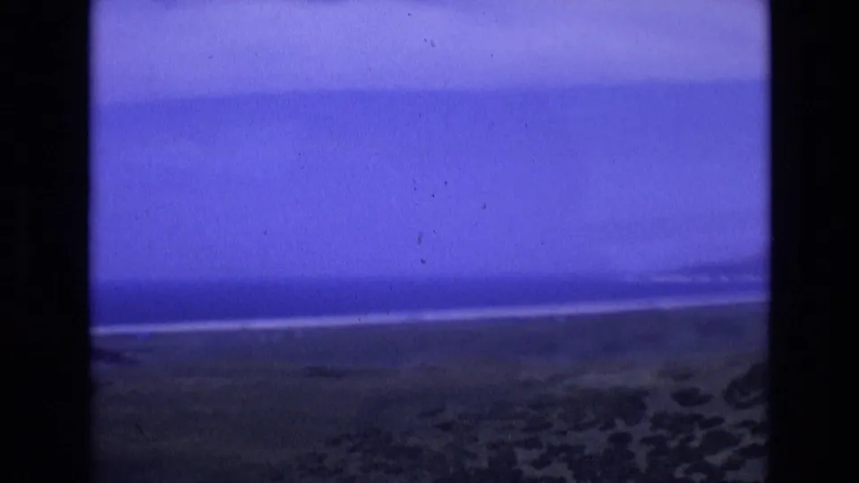 1972 Spain coastal home-movie frame showing a blue twilight sky, thin ocean horizon, sandy beach, and dark shoreline texture within the 8mm frame.