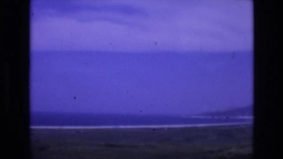 1972 Spain archival frame with a broad ocean band, low coastline, muted foreground terrain, and strong purple color shift from the 8mm film scan.