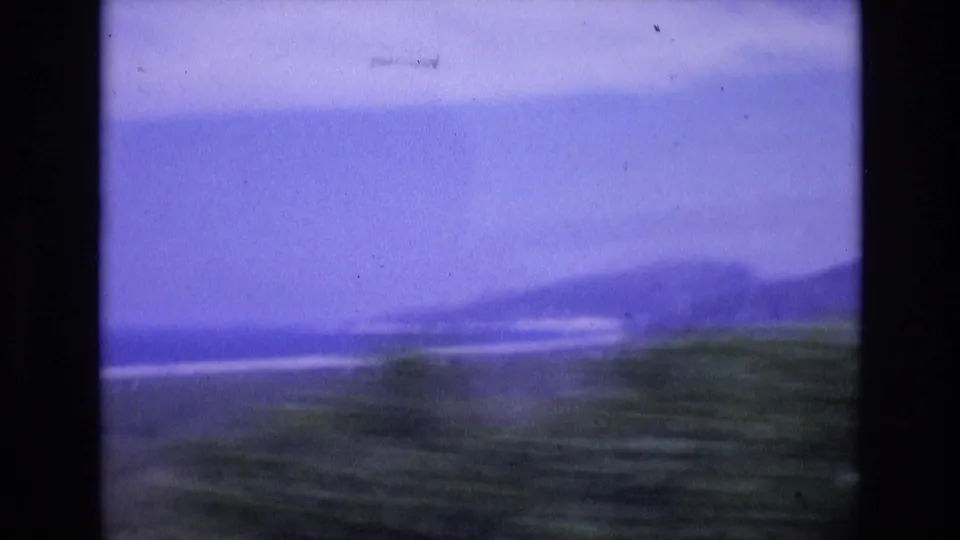 1972 Spain travel frame showing a soft, possibly moving view of distant hills, pale shoreline, ocean water, and green foreground vegetation.