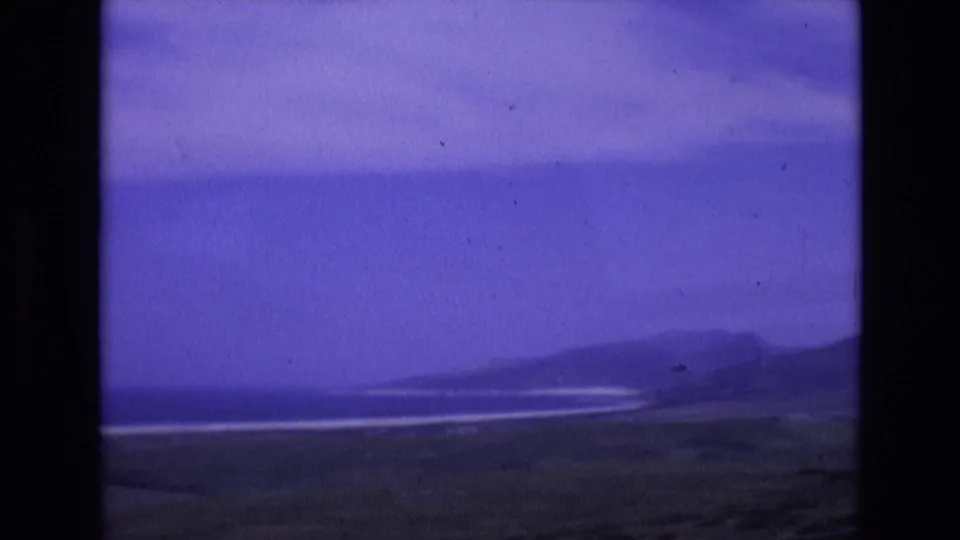 1972 Spain home-movie frame with low hills across the water, bright shoreline bands, dark coastal foreground, and a violet-tinted sky within 8mm frame borders.