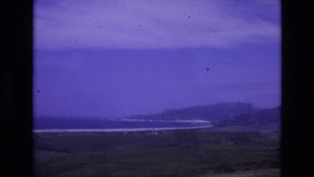 1972 Spain 8mm frame showing a distant coastline with low hills, pale surf bands, dark grassy foreground and a blue-purple sky.