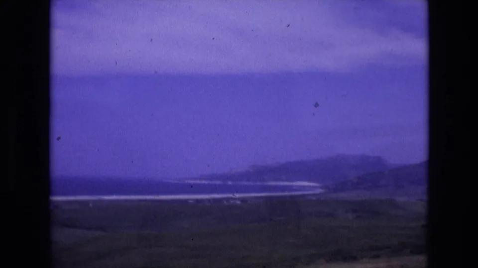 1972 Spain 8mm frame showing a distant coastline with low hills, pale surf bands, dark grassy foreground and a blue-purple sky.
