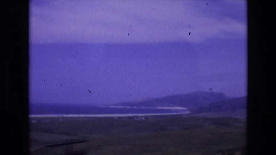 1972 Spain 8mm frame of a wide shoreline and low coastal hills, with muted foreground land and a hazy ocean horizon.