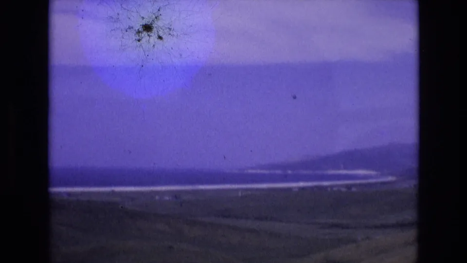 1972 Spain 8mm frame showing pale surf lines below low coastal hills, with dark land in front and visible film dust near the upper sky.