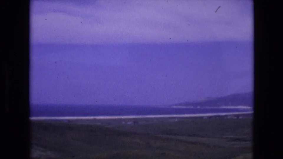 1972 Spain 8mm frame of a soft coastal panorama, with a hazy ocean horizon, pale surf bands and low hills toward the right side of the frame.