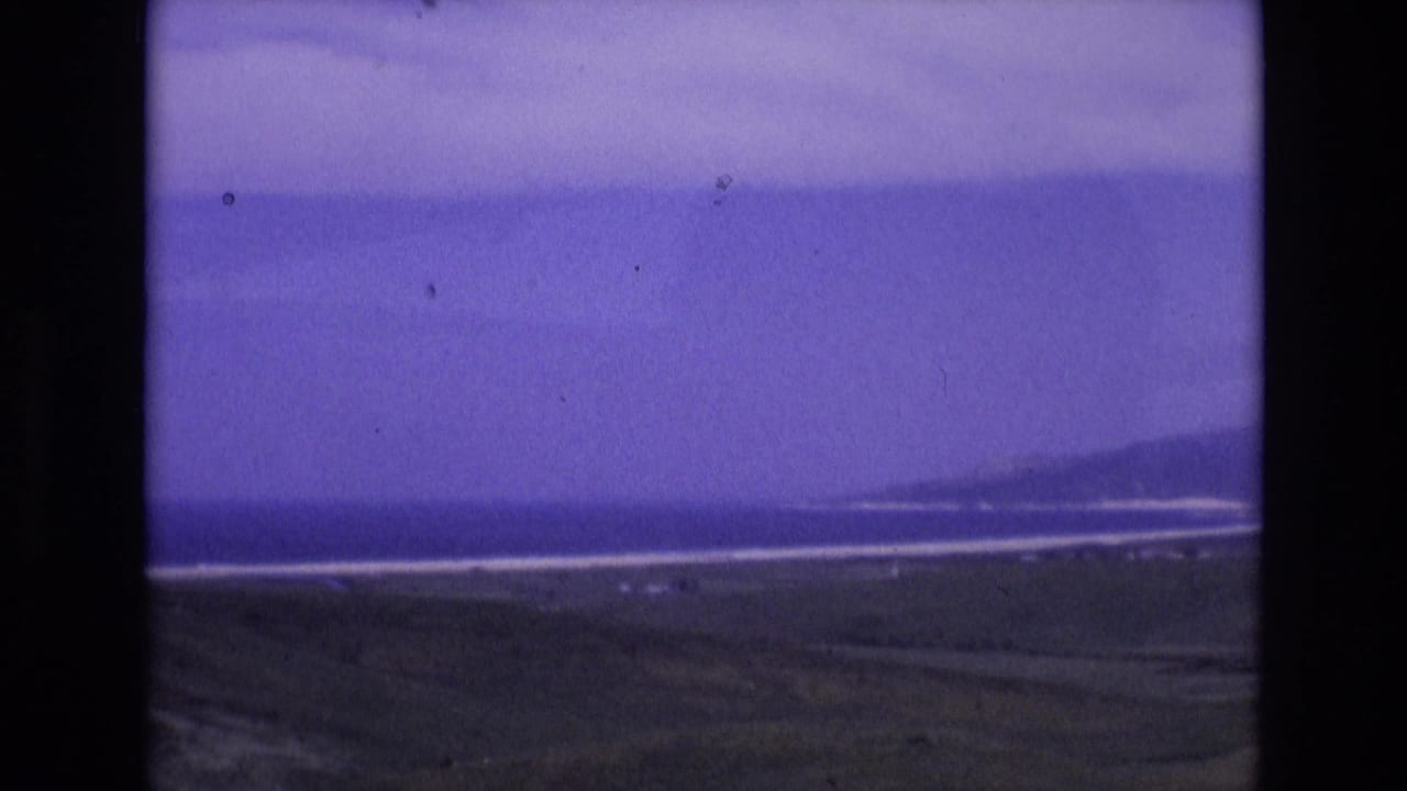 Spain 1972 8mm home-movie frame showing a hazy coastal view with water, pale surf bands, low land, and distant hills under a color-shifted sky.