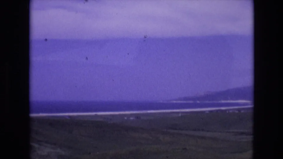 Spain 1972 8mm home-movie frame showing a hazy coastal view with water, pale surf bands, low land, and distant hills under a color-shifted sky.