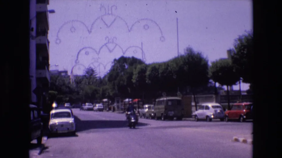 Spain 1972 street scene with parked 1970s compact cars, a tree-lined road, distant pedestrians or riders, and a decorative overhead street arch.