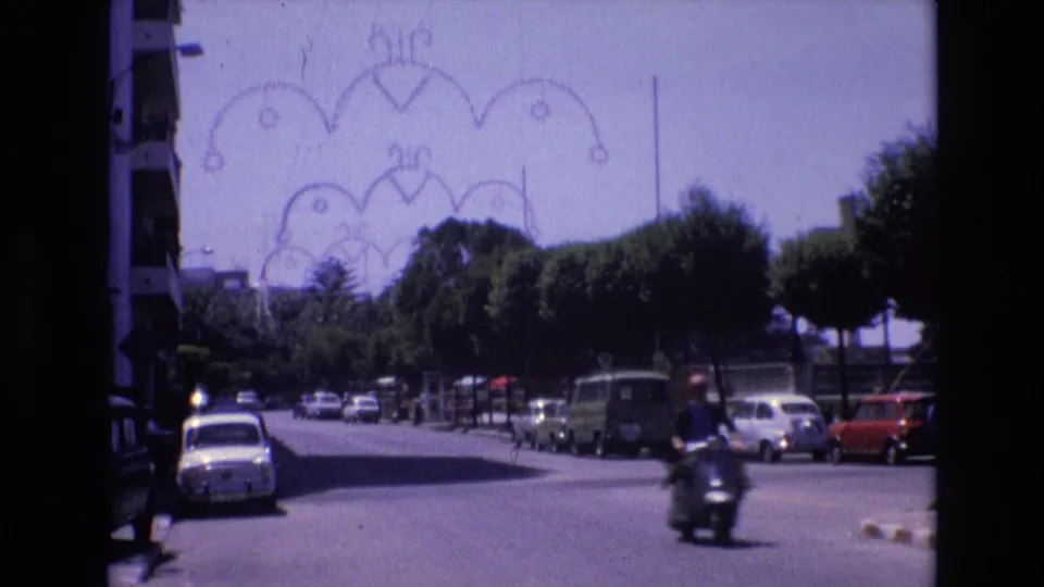 Spain 1972 urban home-movie frame showing a motorbike moving through a sunlit street lined with parked compact cars and trees beneath a decorative arch.