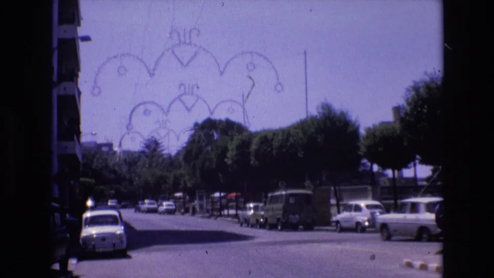 Spain 1972 8mm frame of a sunlit urban road with parked cars along both sides, clipped trees, and a decorative arch structure in the background.