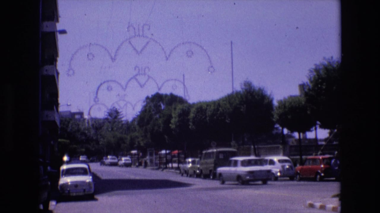 1972 Spain home-movie frame showing a sunlit tree-lined street near the coast with parked cars and decorative overhead street fixtures.