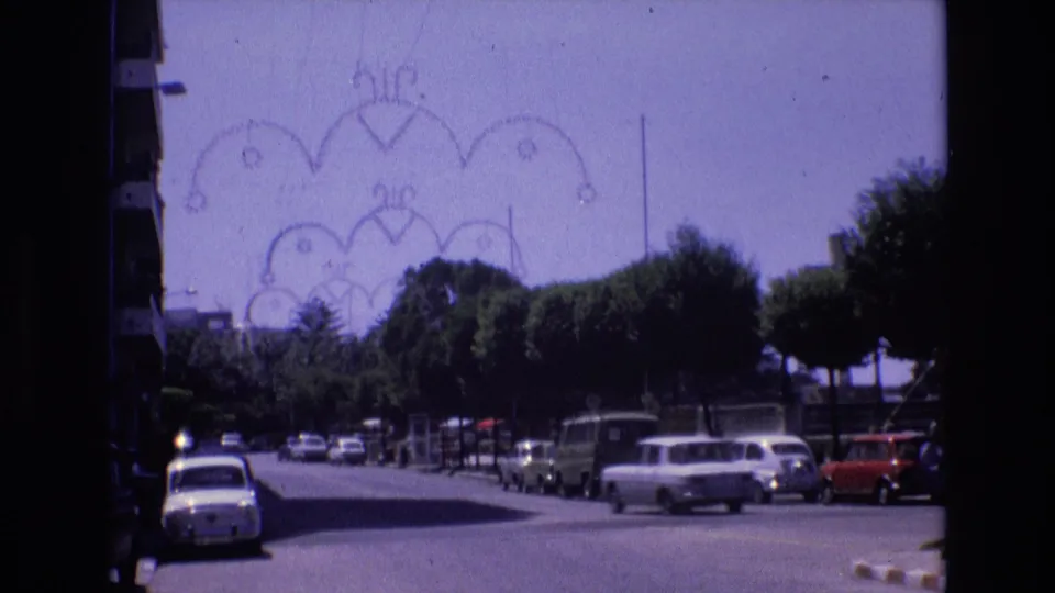 1972 Spain home-movie frame showing a sunlit tree-lined street near the coast with parked cars and decorative overhead street fixtures.