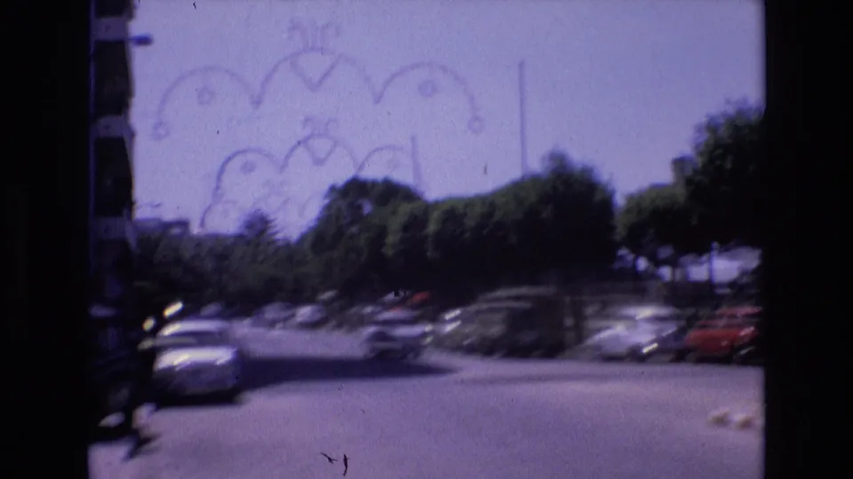 Soft, handheld 1972 Spain frame showing a moving view along a sunlit street with trees, parked cars, and decorative overhead fixtures.