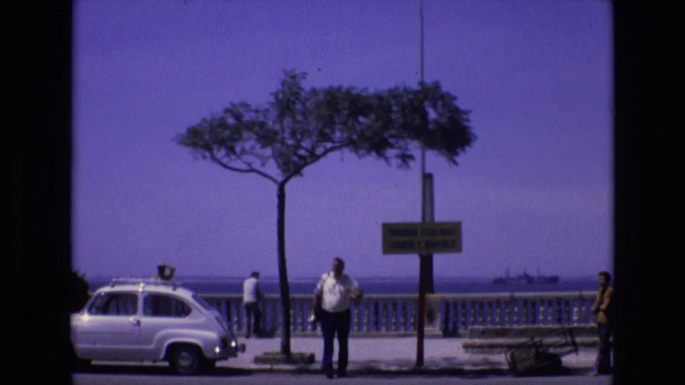 1972 Spain home-movie frame showing a coastal promenade with a white compact car, a single tree, a yellow sign, pedestrians, the sea, and a distant ship.