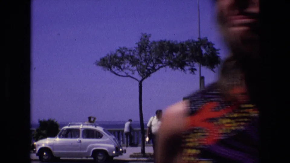 1972 Spain home-movie frame showing the coastal promenade with a white compact car, pedestrians near the railing, open sea, a tree, and a blurred woman passing close to camera.