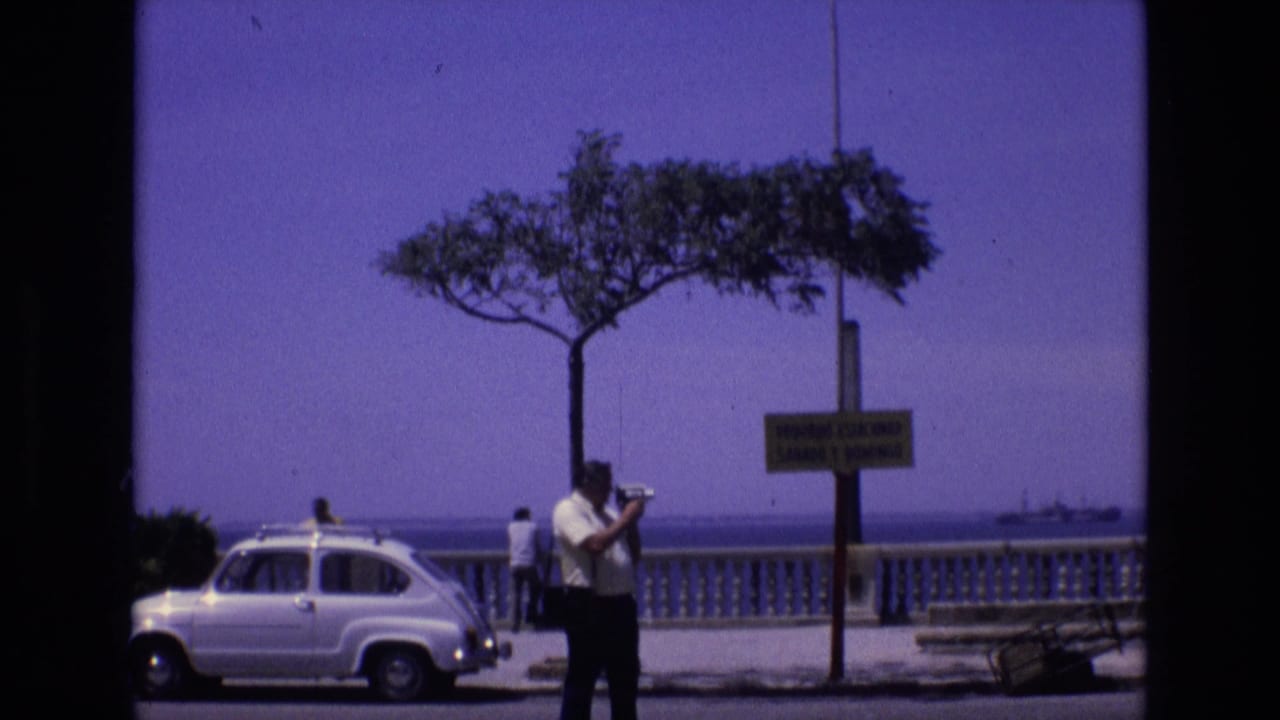In Spain in 1972, a man with a camera stands on a coastal promenade beside a white compact car, with a tree, signpost, balustrade, and sea in the background.