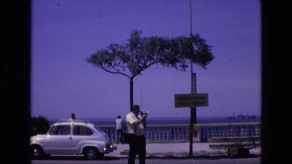 In Spain in 1972, a man with a camera stands on a coastal promenade beside a white compact car, with a tree, signpost, balustrade, and sea in the background.