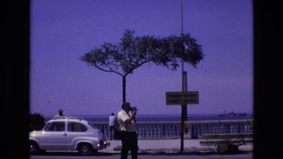 A 1972 Spanish coastal promenade view shows a man taking a photograph near a white vintage car, with a wind-shaped tree, signpost, balustrade, and sea beyond.