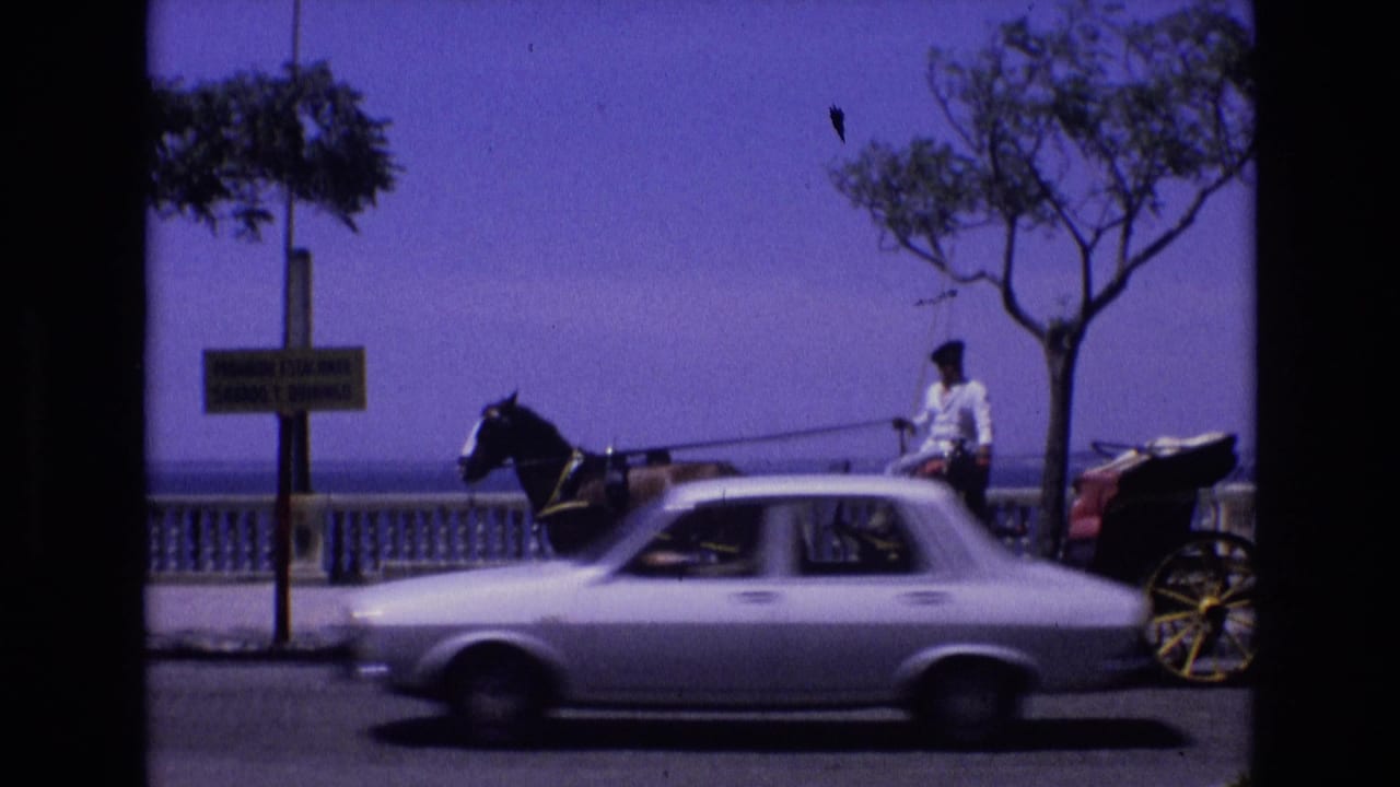 Spain 1972 8mm frame showing a horse-drawn buggy and seated driver near a seaside promenade while a white car moves past in the foreground.