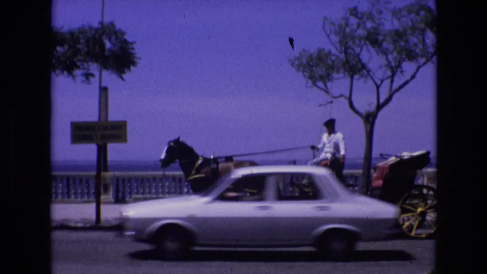 Spain 1972 8mm frame showing a horse-drawn buggy and seated driver near a seaside promenade while a white car moves past in the foreground.