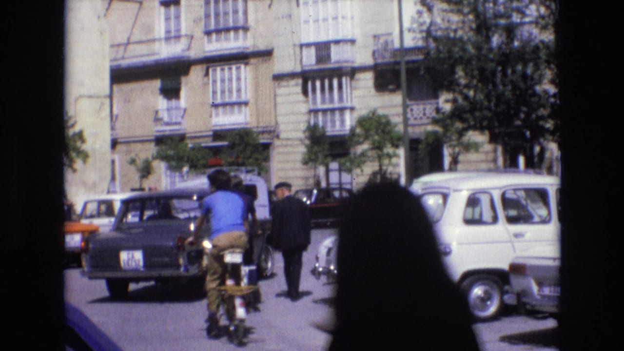 In 1972 Spain, a cyclist moves through traffic on a sunlit old-town street lined with compact cars and balconied apartment buildings.