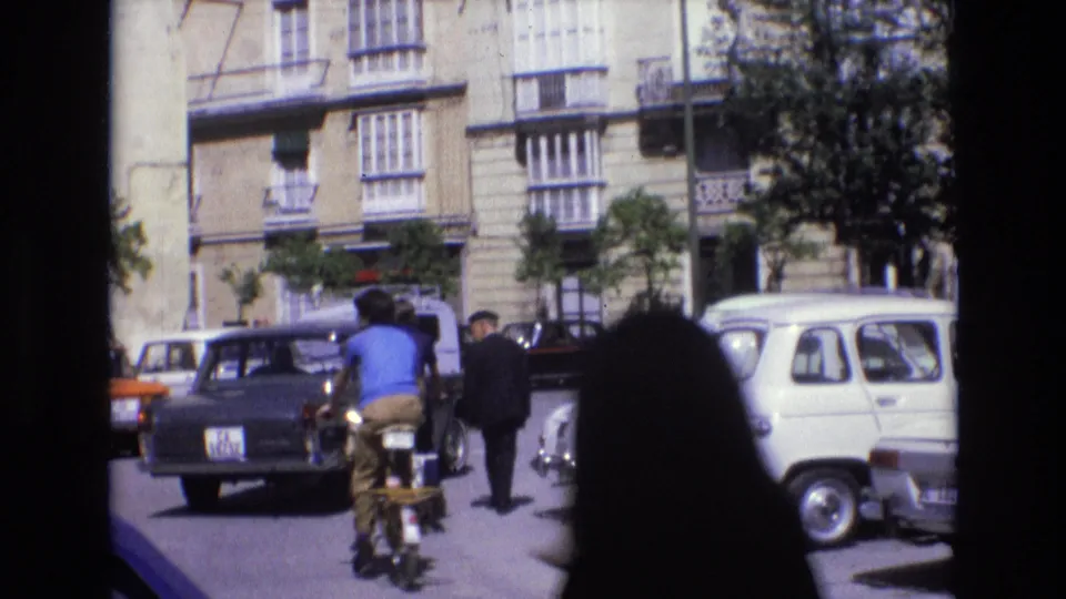 In 1972 Spain, a cyclist moves through traffic on a sunlit old-town street lined with compact cars and balconied apartment buildings.