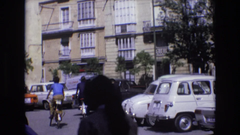 A cyclist travels through a Spanish street in 1972, passing compact cars and balconied buildings while a dark foreground silhouette partly frames the view.