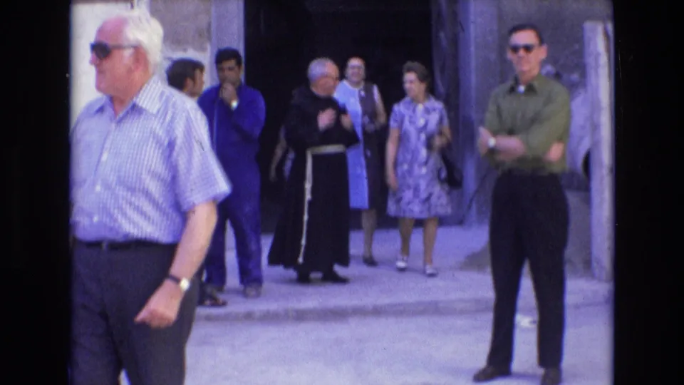Adults in 1970s clothing gather near a stone entrance in Spain, with a dark-robed cleric among pedestrians and a man in sunglasses standing at the edge of the frame.