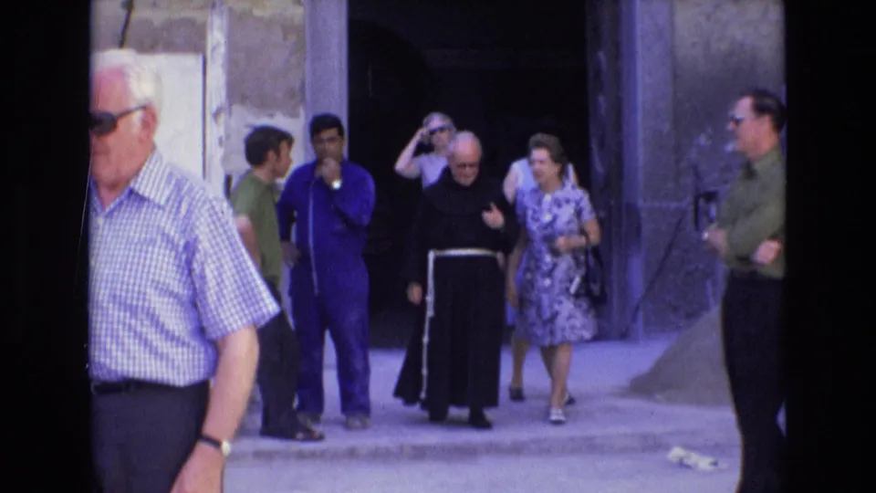 A group of adults stands outside a stone old-town entrance in 1972 Spain, including a dark-robed cleric, a woman in a patterned dress, and men wearing sunglasses.
