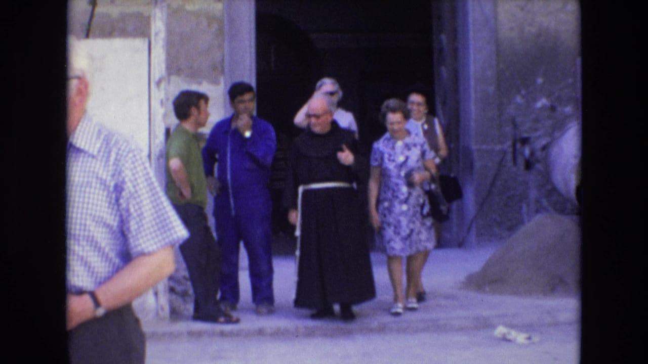 In Spain in 1972, a monk in a black habit with a white cord walks with casually dressed tourists near a worn plaster wall and dark entrance.