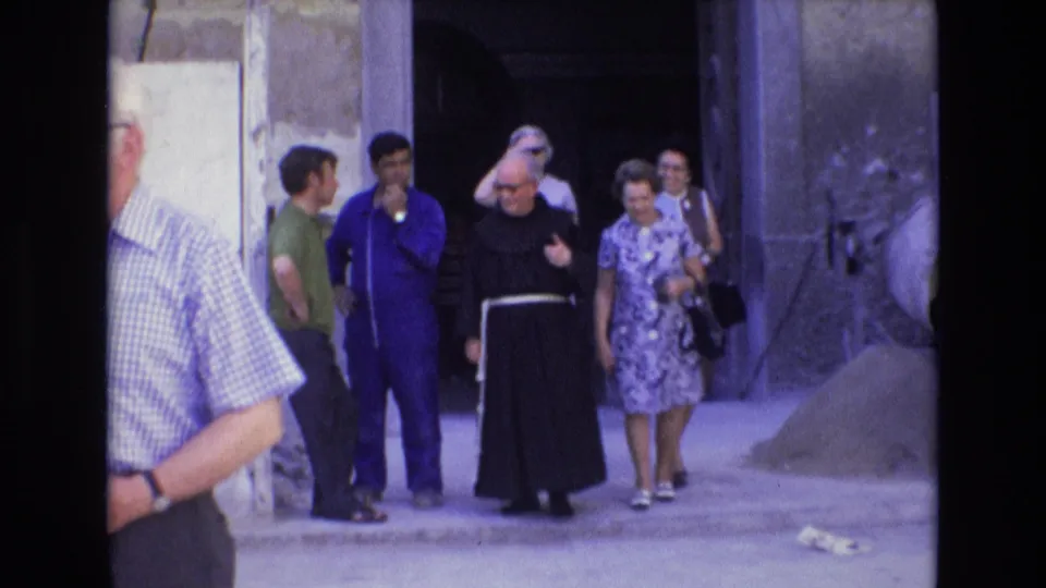 In Spain in 1972, a monk in a black habit with a white cord walks with casually dressed tourists near a worn plaster wall and dark entrance.