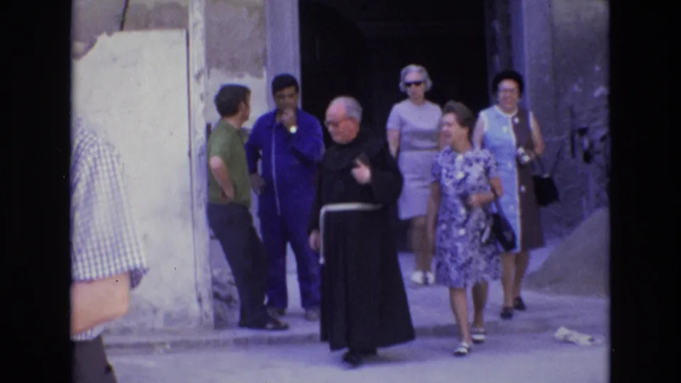 A monk and several adults in 1970s clothing gather near a dark doorway in a narrow Spanish street, with a woman in a patterned dress moving forward.