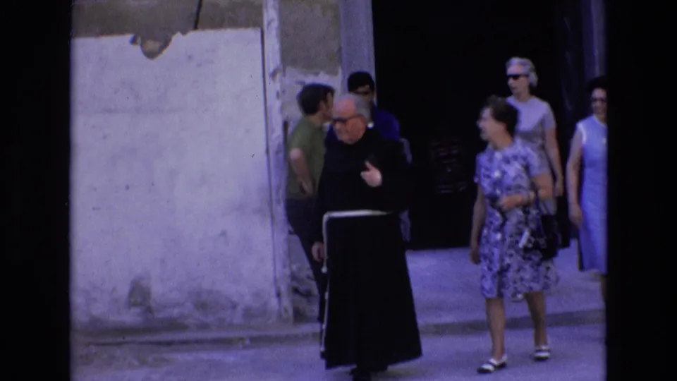 The monk walks beside a woman in a patterned dress as other tourists stand near the entrance behind them in the 1972 Spain home-movie scene.