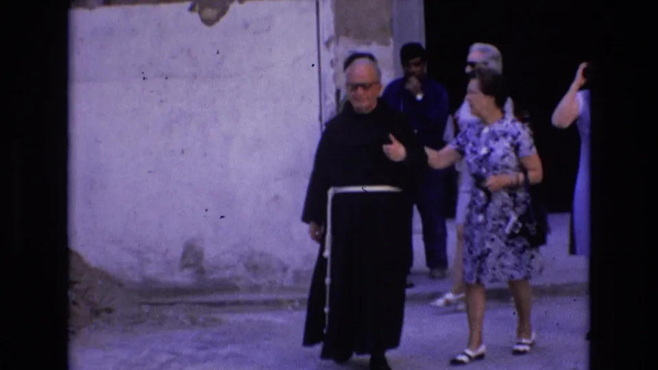 A monk in a black habit and a woman in a patterned dress walk side by side while tourists emerge behind them in a narrow Spanish street.