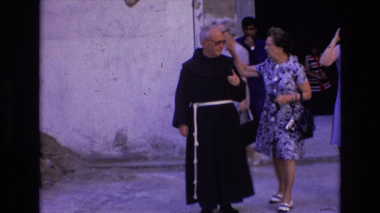 In this 1972 Spain 8mm frame, a priest in a black habit with a white cord walks beside a tourist in a patterned dress near a doorway and pale street wall.