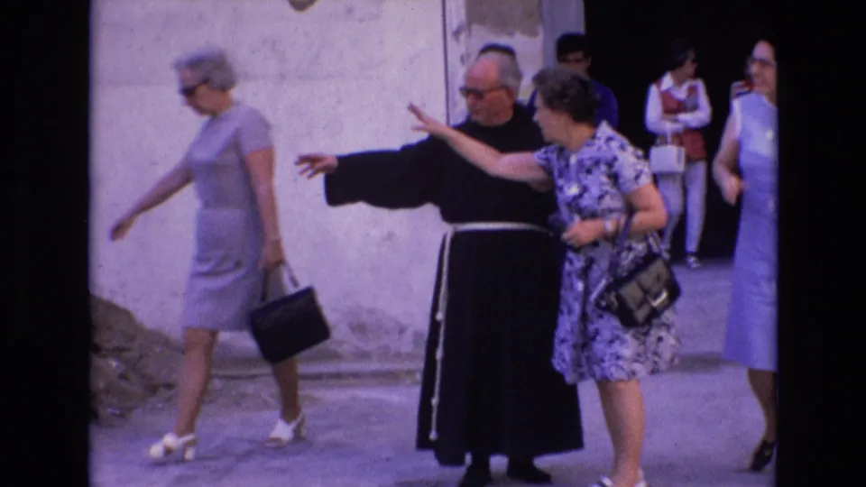 In this Spain 1972 street frame, the priest extends an arm as if indicating a direction while tourists and pedestrians pass near a pale building facade.