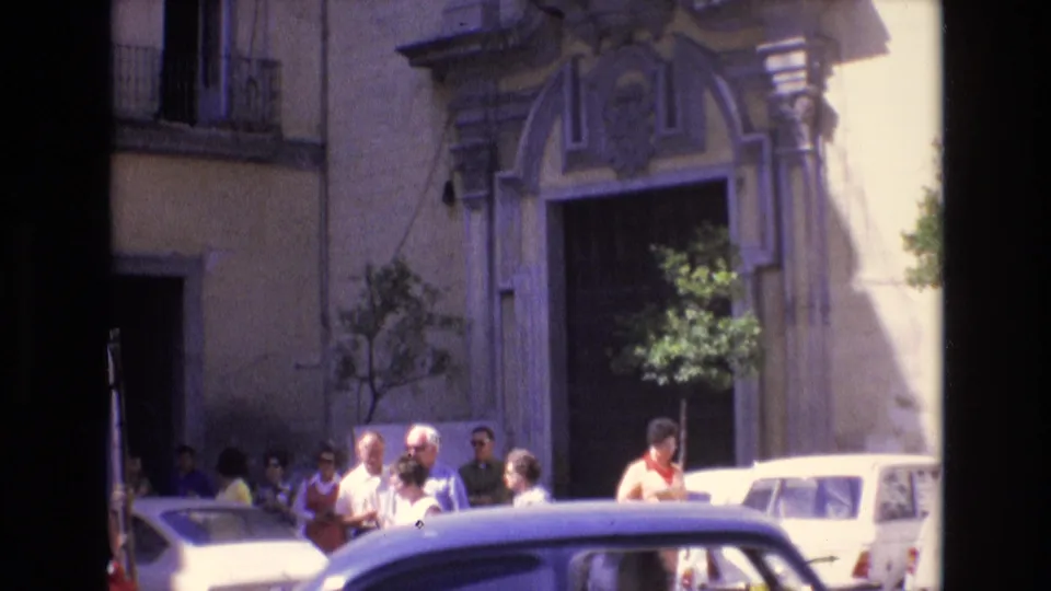 This 1972 Spain 8mm frame shows people gathered near parked period cars and an ornate arched doorway on a sunlit historic street.
