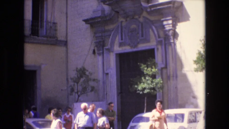 In the closing Spain 1972 frame, a woman stands near parked cars and pedestrians below an ornate arched entrance, with strong sunlight across the facade.