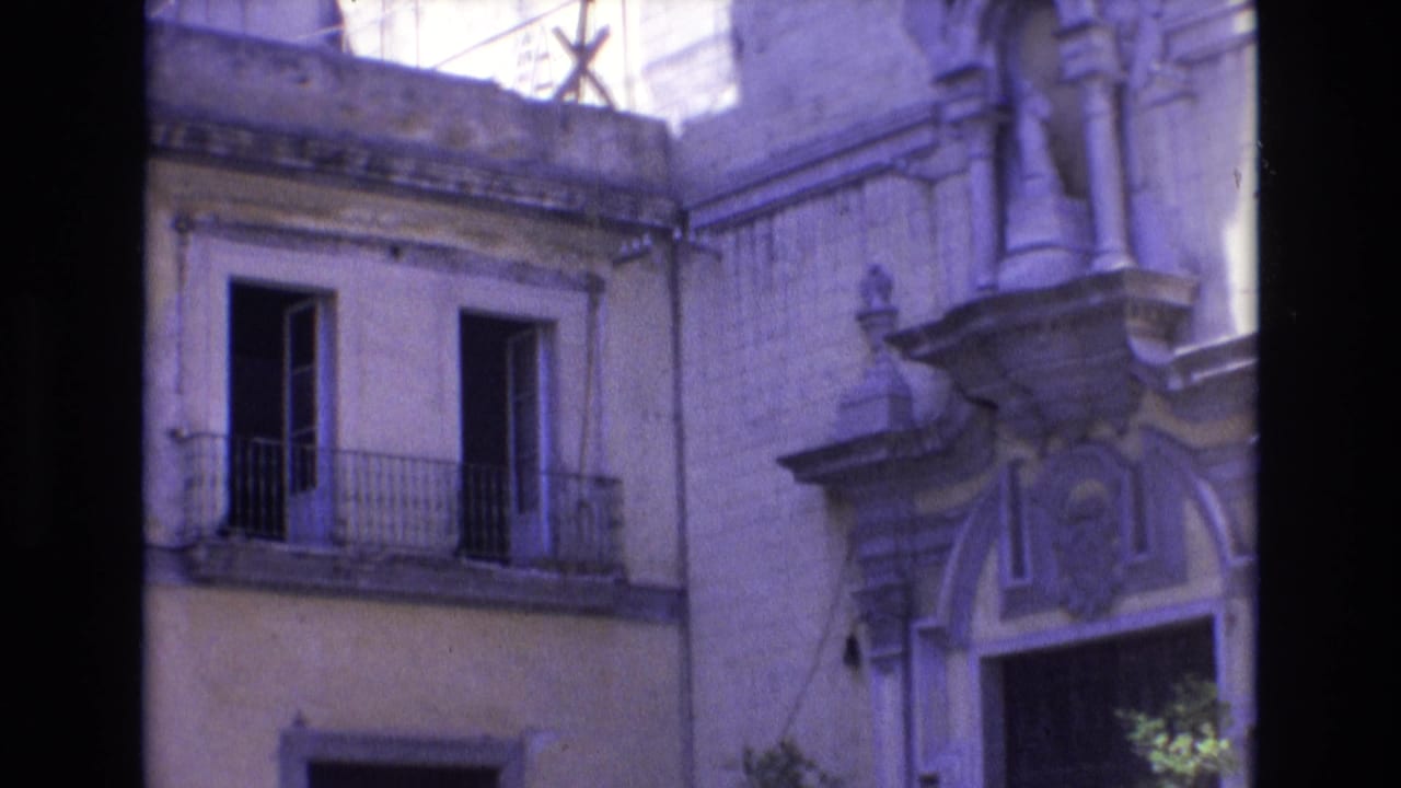 1972 Spain home-movie frame showing a weathered yellow facade with open upper windows, iron balcony railings, and a decorative stone entrance.