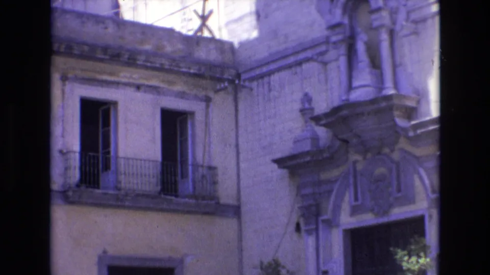 1972 Spain home-movie frame showing a weathered yellow facade with open upper windows, iron balcony railings, and a decorative stone entrance.
