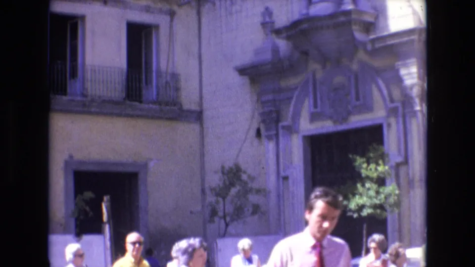 1972 Spain home-movie frame with pedestrians in summer clothing passing in front of an ornate historic building entrance and balcony.