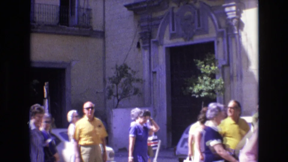 1972 Spain street frame showing several adults walking or standing near an ornate building entrance, with a partial light-colored car and greenery in the background.