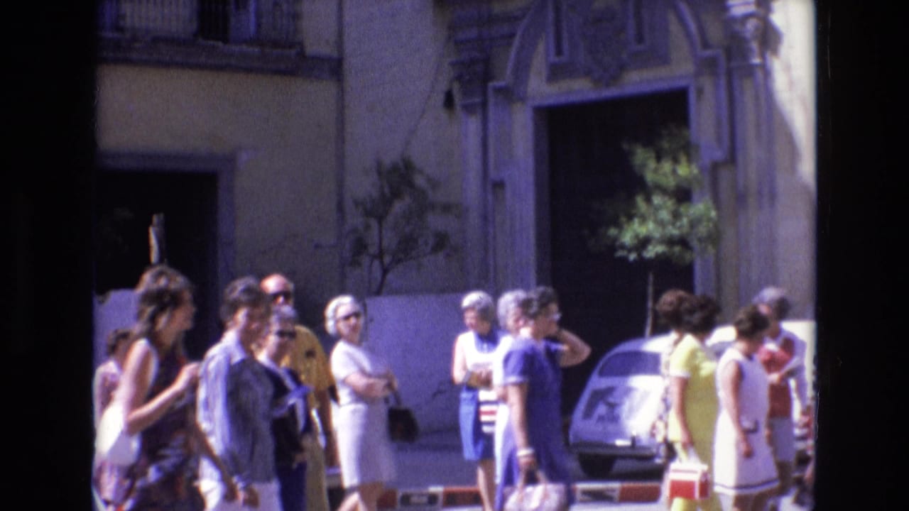 1972 Spain home-movie frame showing pedestrians in summer clothing near a small car and an ornate sunlit building facade on a town street.
