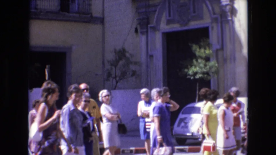 1972 Spain home-movie frame showing pedestrians in summer clothing near a small car and an ornate sunlit building facade on a town street.