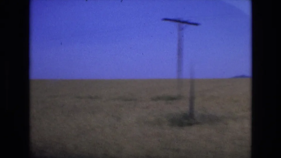 1972 Spain home-movie travel frame with a roadside pole crossing a grassy field and blue sky, suggesting movement past rural terrain.