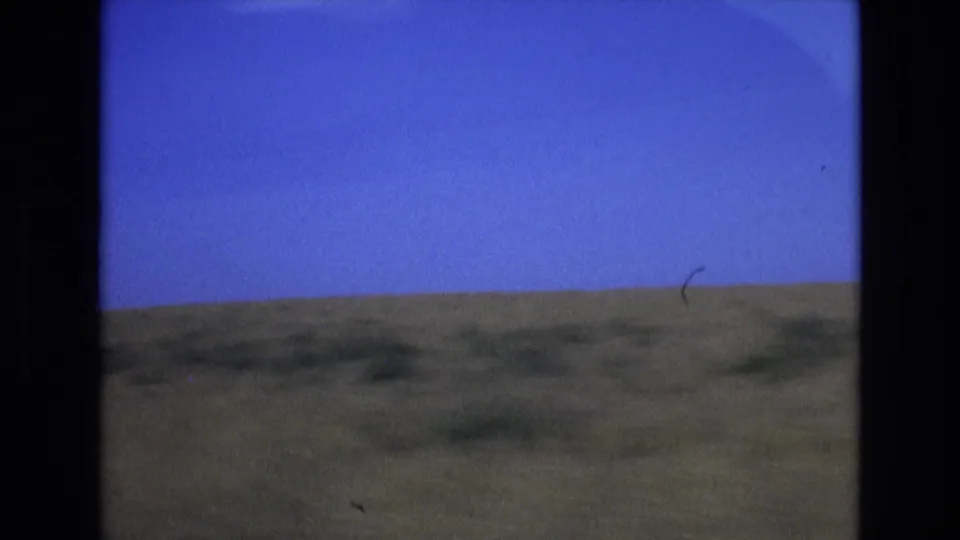 1972 Spain 8mm frame with a broad blue sky over a muted green-brown plain, bordered by the scanned film gate and marked by small dust artifacts.