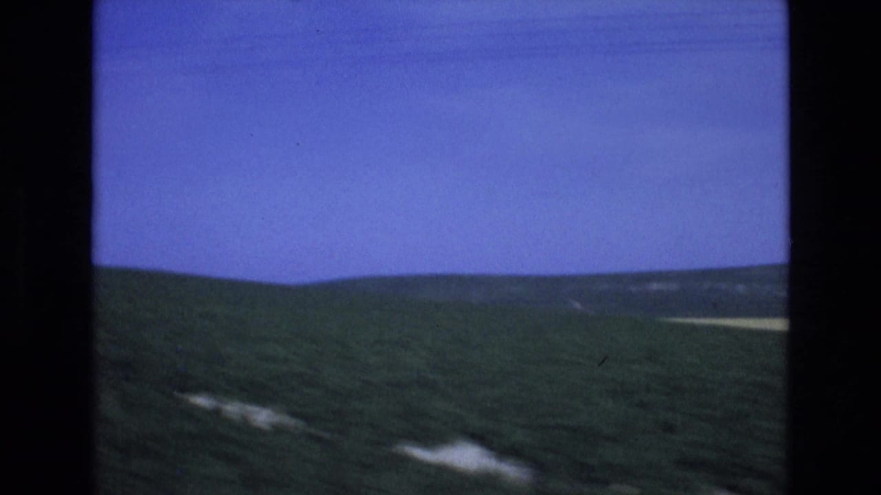 1972 Spain 8mm frame showing rolling green hills under a saturated blue sky, viewed from inside a moving car with dark window edges.