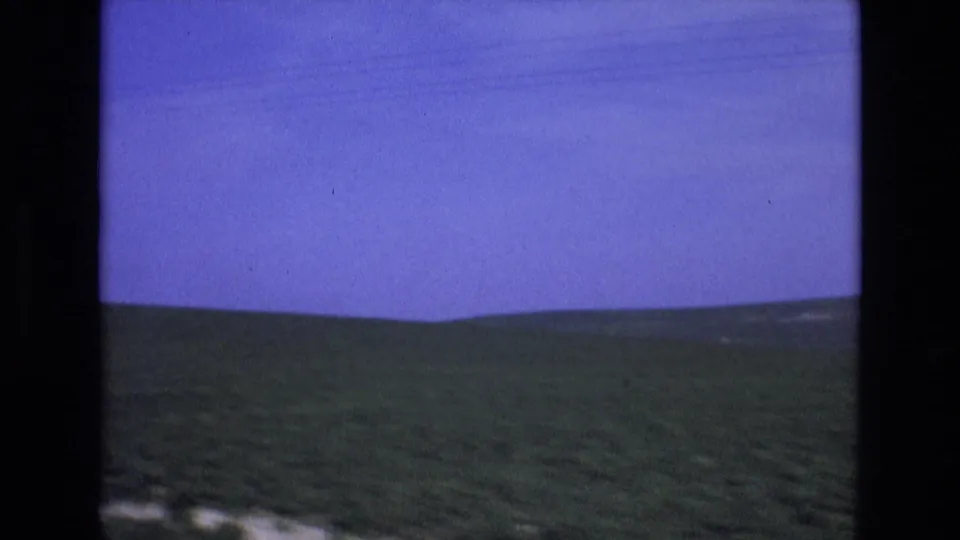 1972 Spain 8mm still with a blurred rural hillside, saturated sky, and dark car-window edge indicating movement along a road.