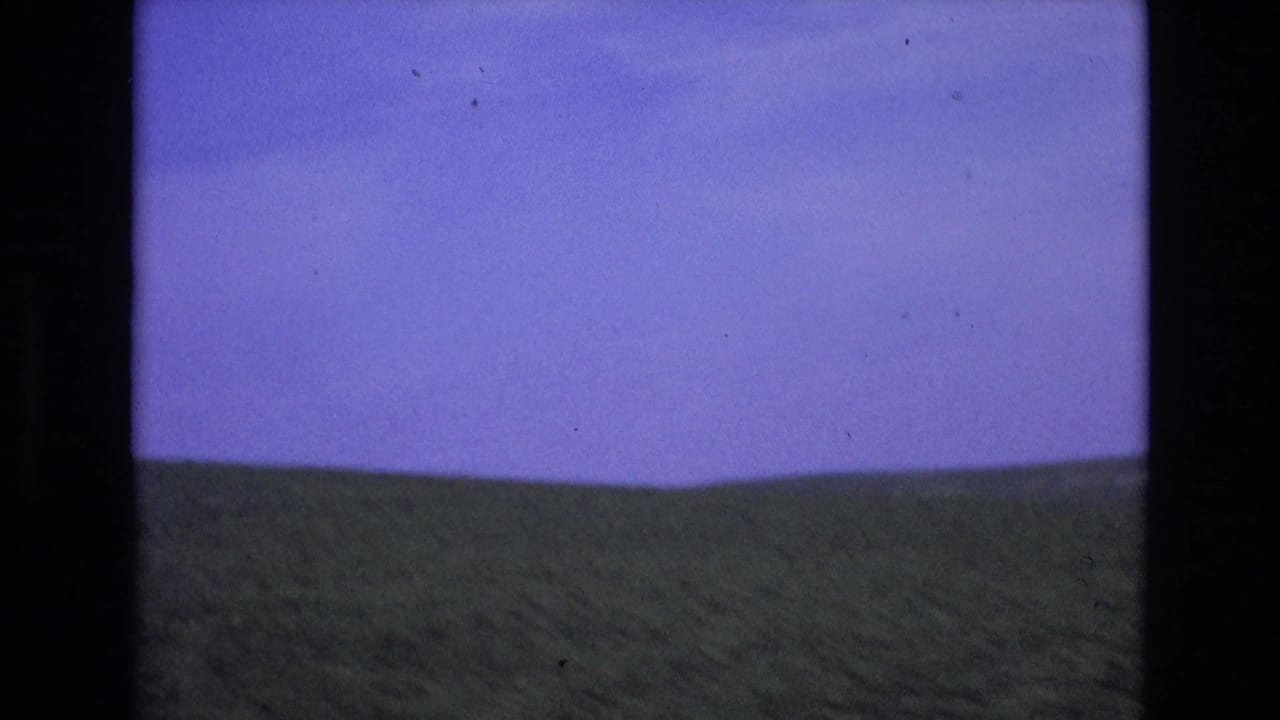 Metadata-supported 1972 Spain home-movie frame showing a green valley and low hills beneath a saturated blue sky, framed by dark 8mm gate edges.